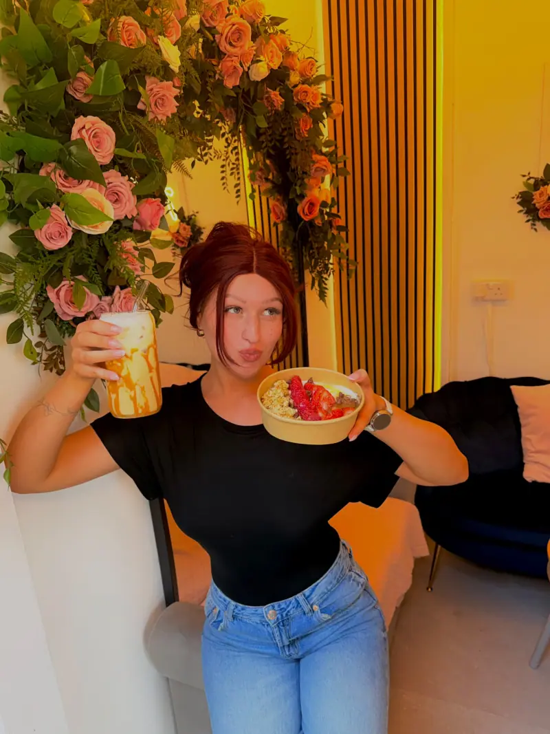 Lucy holding an iced drink and a strawberry-topped bowl beside Tina Rose's floral wall