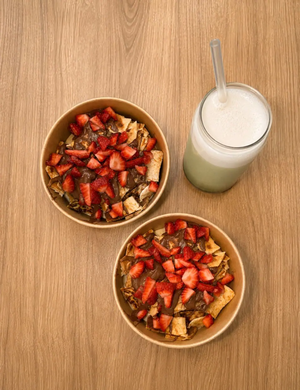 Two strawberry and chocolate crepe bowls with a frothy green drink on a wooden table